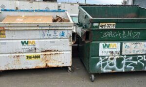 Two large industrial dumpsters, one heavily rusted and the other covered in graffiti, placed near a wall.
